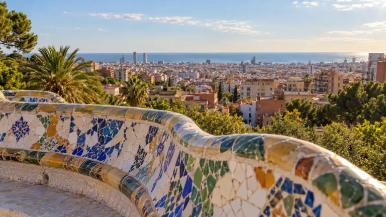 Home 23 AI-generated view of a curved mosaic bench at Park Güell with Barcelona rooftops and the Mediterranean Sea beyond.